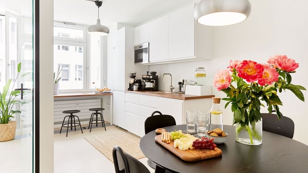 Blick in eine Büro-Küche mit weisser Küchenzeile und einem schwarzen, runden Esstisch, auf dem Blumen und Snacks stehen. 