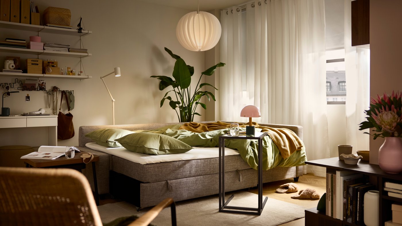 A FRIHETEN sofa bed in bed mode with its bedding undone, in a softly lit living room with curtains and blinds mostly shut.