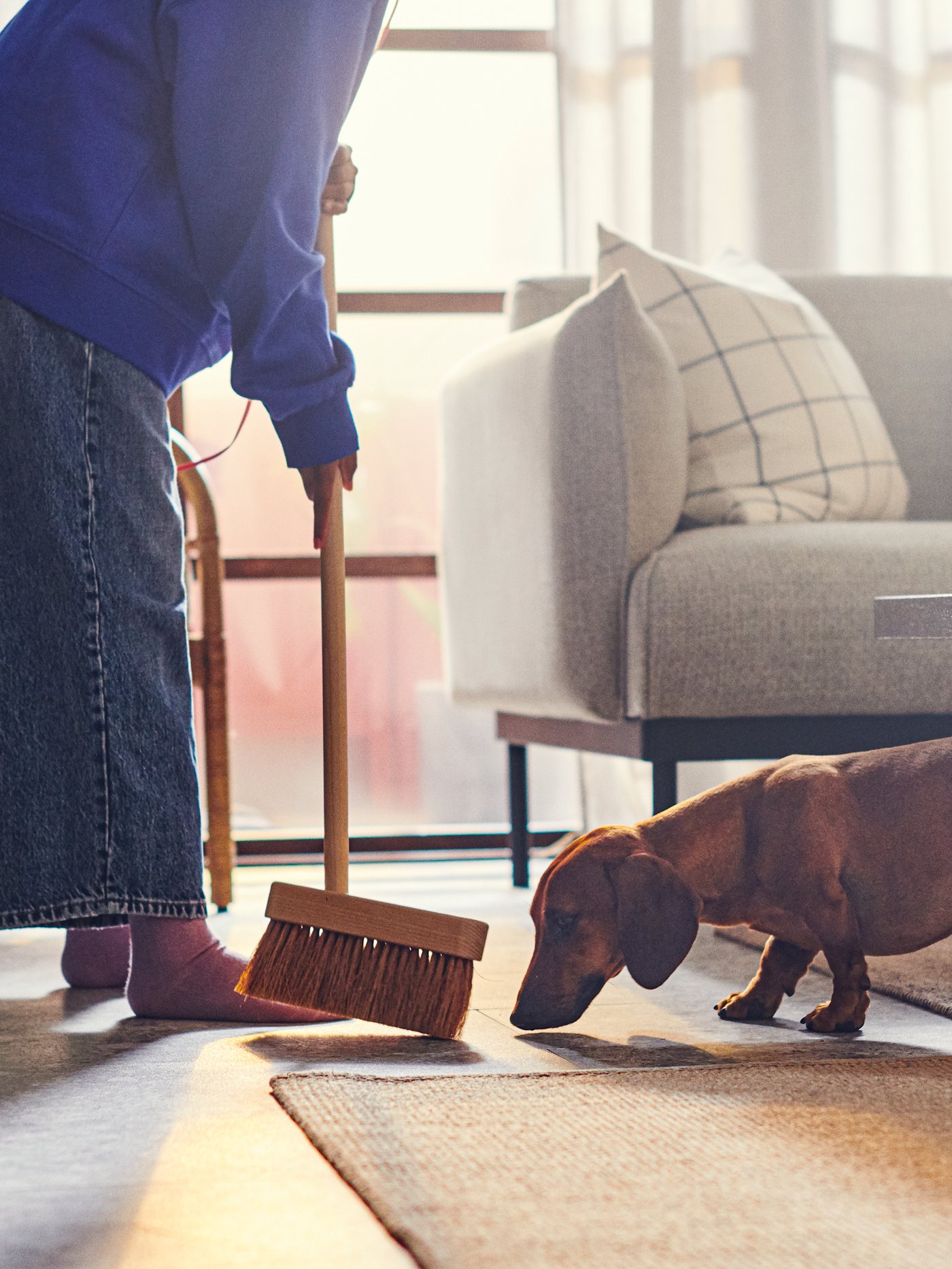 A person is sweeping the floor with a broom while a dog looks on.