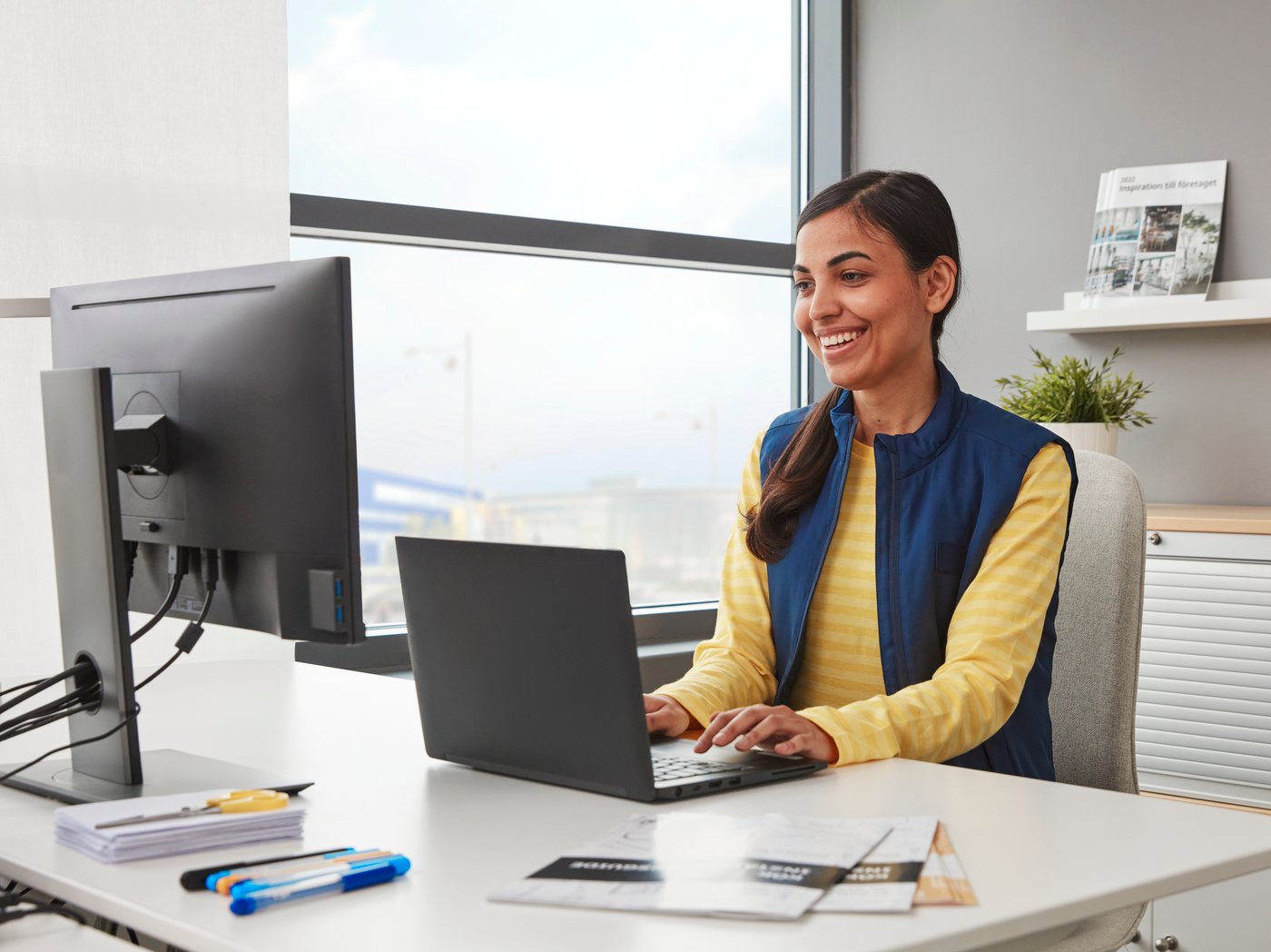 L'image montre une personne assise à un bureau, travaillant sur un ordinateur portable, avec un écran, des papiers, des stylos et un magazine à côté, tandis qu'une fenêtre et des étagères sont visibles à l'arrière-plan.