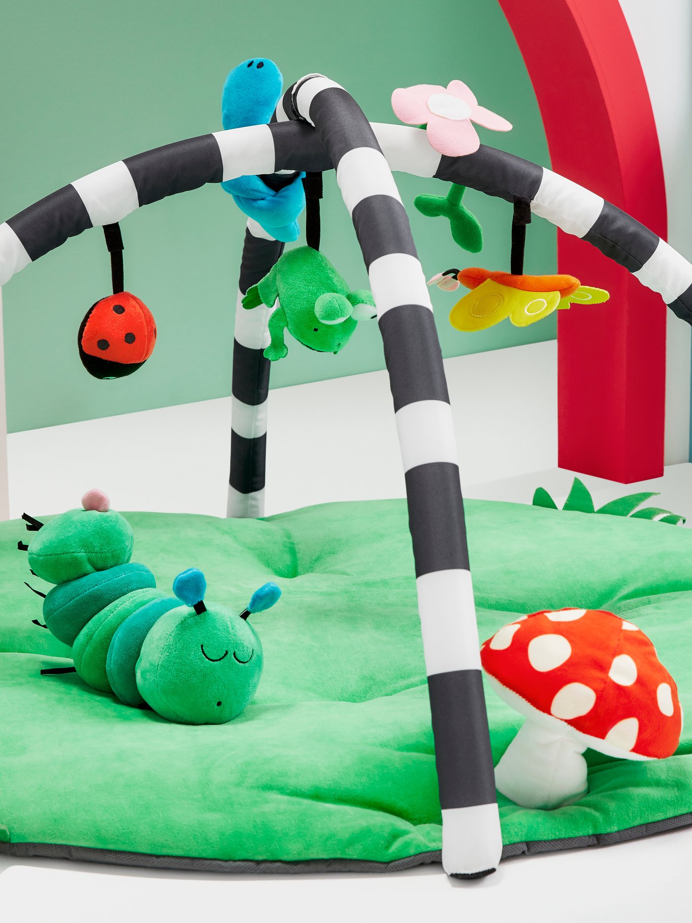 Un gimnasio para bebés KLAPPA de colores verde, blanco y negro con figuras de hongos, flores y animales. Sobre el gimnasio hay un sonajero KLAPPA en forma de oruga verde. 
