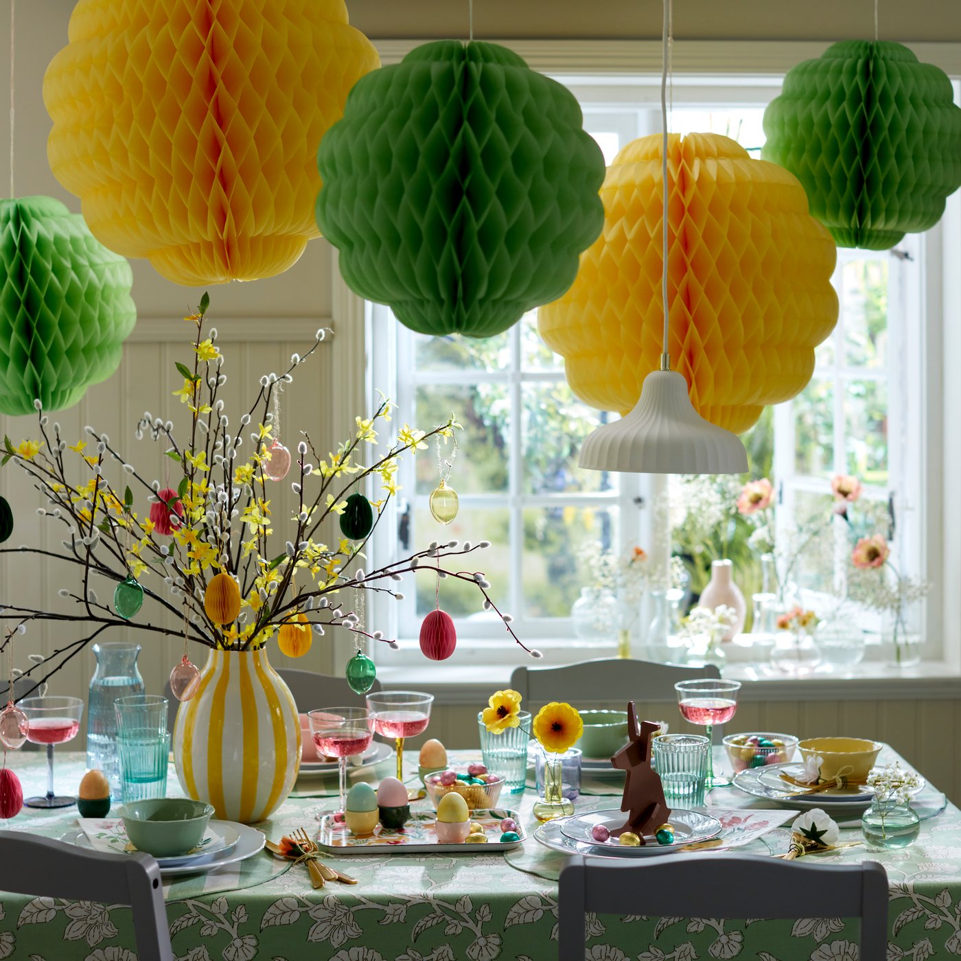 Een vrolijke eettafel met gekleurd TJÄRLEK-glaswerk, omringd door feestelijke TJÄRLEK-hangdecoraties.