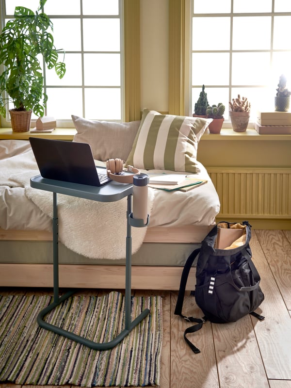 A turquoise laptop stand is placed by a bed that functions as a seat. A grey backpack is placed next to the bed.