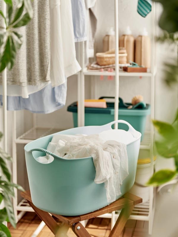 Wet textiles in a light blue TORKIS laundry basket set on a stool next to garments drying on a white JOSTEIN shelving unit.