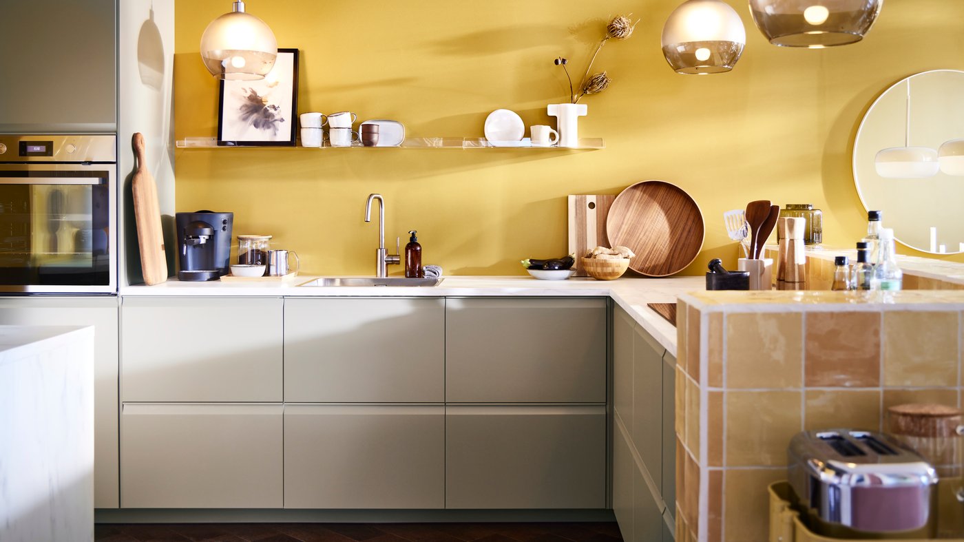 L-Form Küche mit UPPLÖV Schubladenfronten in mattem Dunkelbeige, Hängeleuchten mit JAKOBSBYN Hängeleuchtenschirmen in Frostglas und Grau und andere Küchengegenständen.