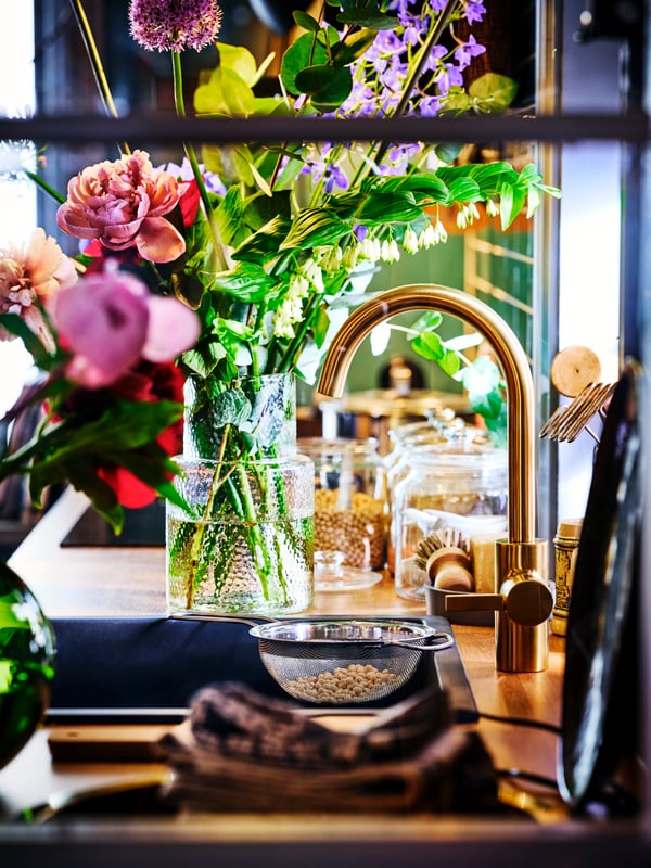 A kitchen area with a DELSJÖN kitchen mixer tap in brass color, with a colorful flower arrangement in a glass vase behind.