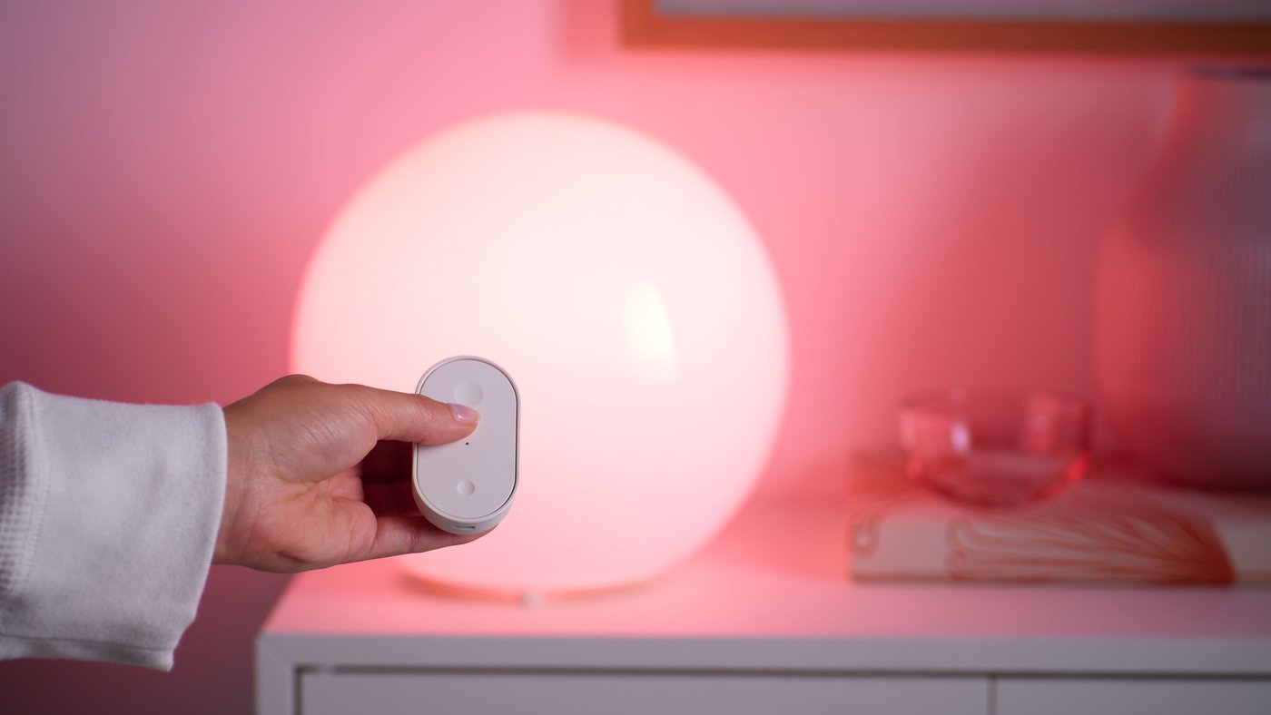 Hand håller en vit fjärrkontroll framför en rund smart bordslampa med varmt rosa sken på en vit byrå.