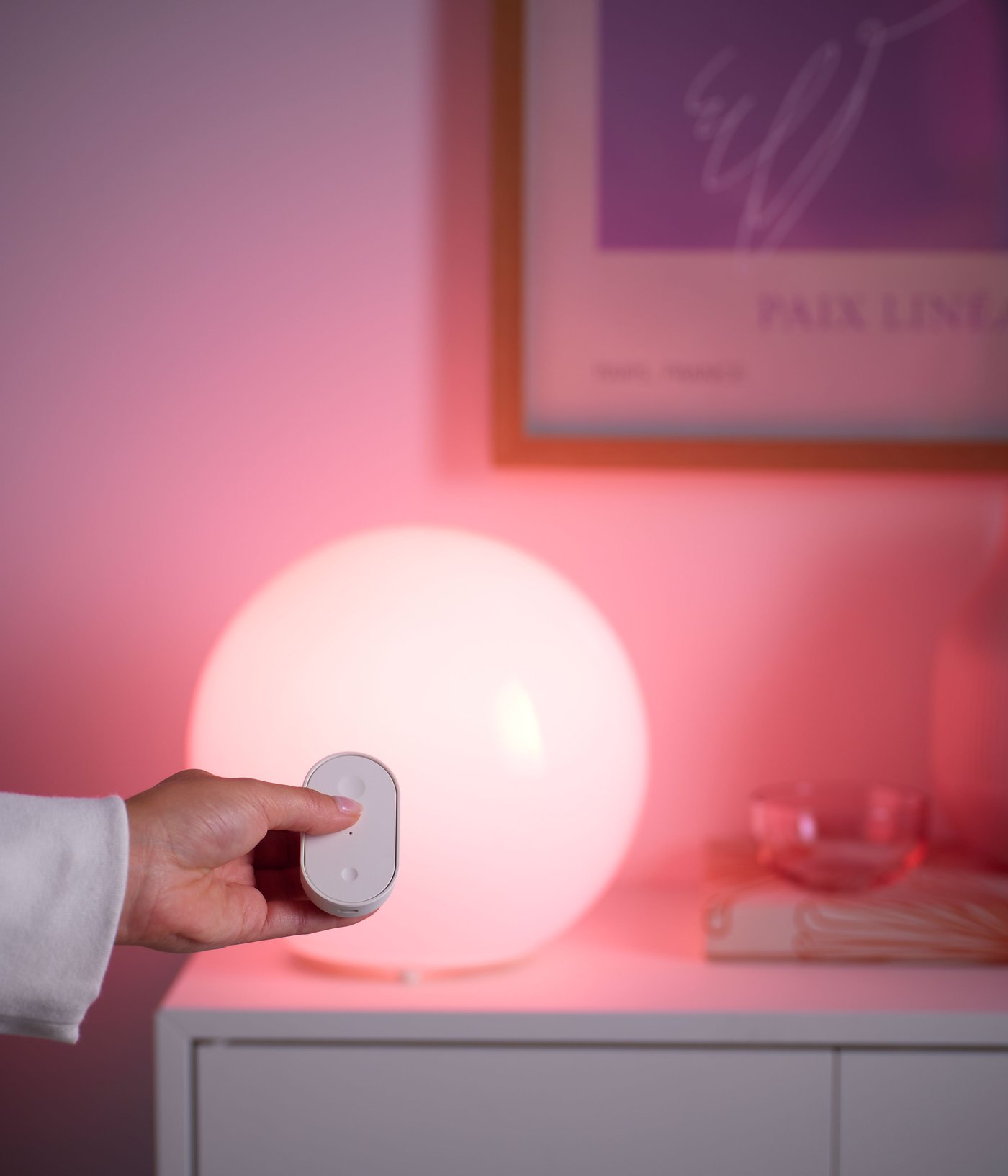 Hand holding a white remote control beside a round smart table lamp glowing warm pink on a white cabinet.