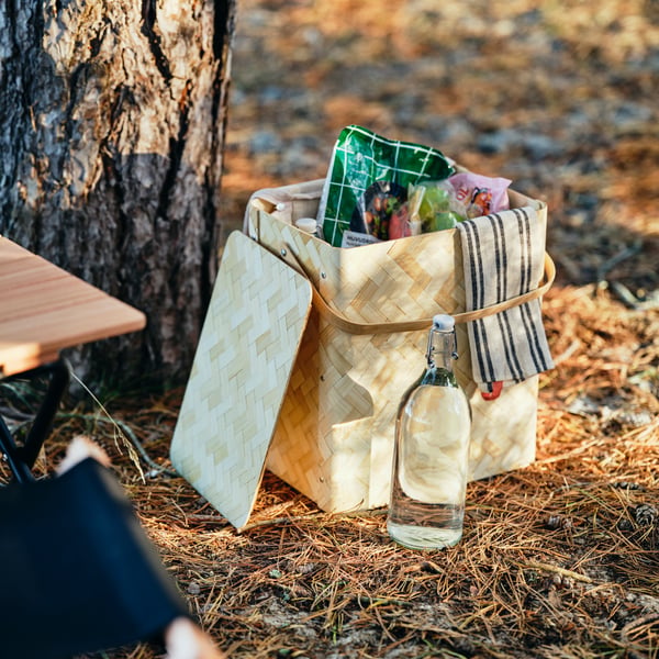 A SOLUPPGÅNG cool basket with a pack of HUVUDROLL plant balls and other packed food items in it stands next to a tree.