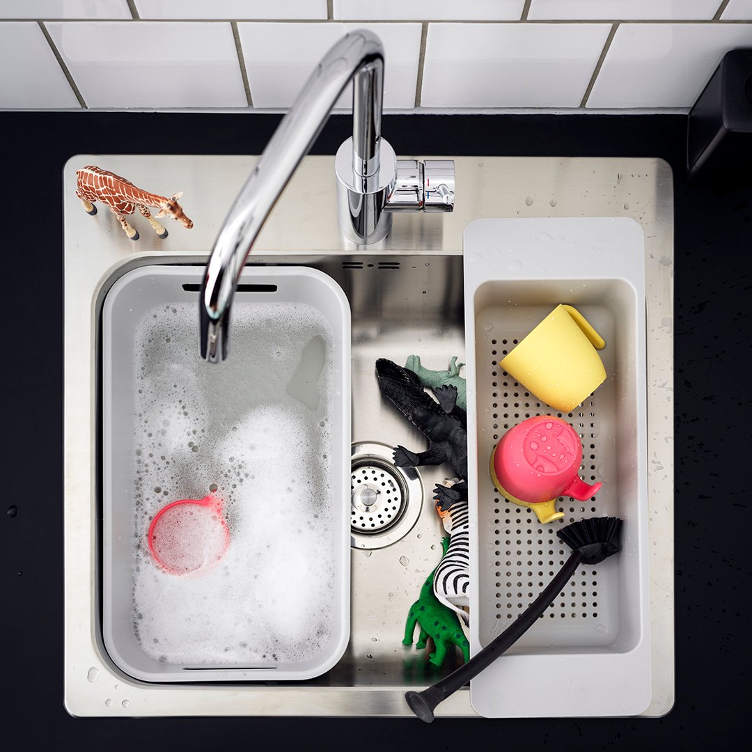La imagen muestra un lavaplatos blanco visto desde arriba. La llave de agua cromada apunta a un recipiente donde se acumula el agua y el jabón para lavar los platos. Al lado derecho hay una rejilla con dos tazas y un cucharón. Bajo los dos recipientes se ven animales de juguete y la rejilla que protege el desagüe de los desperdicios.
