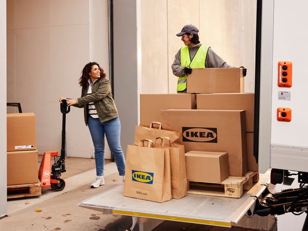 A smiling customer holding a pallet jack with large IKEA packages, talking to the driver who has more packages to offload.