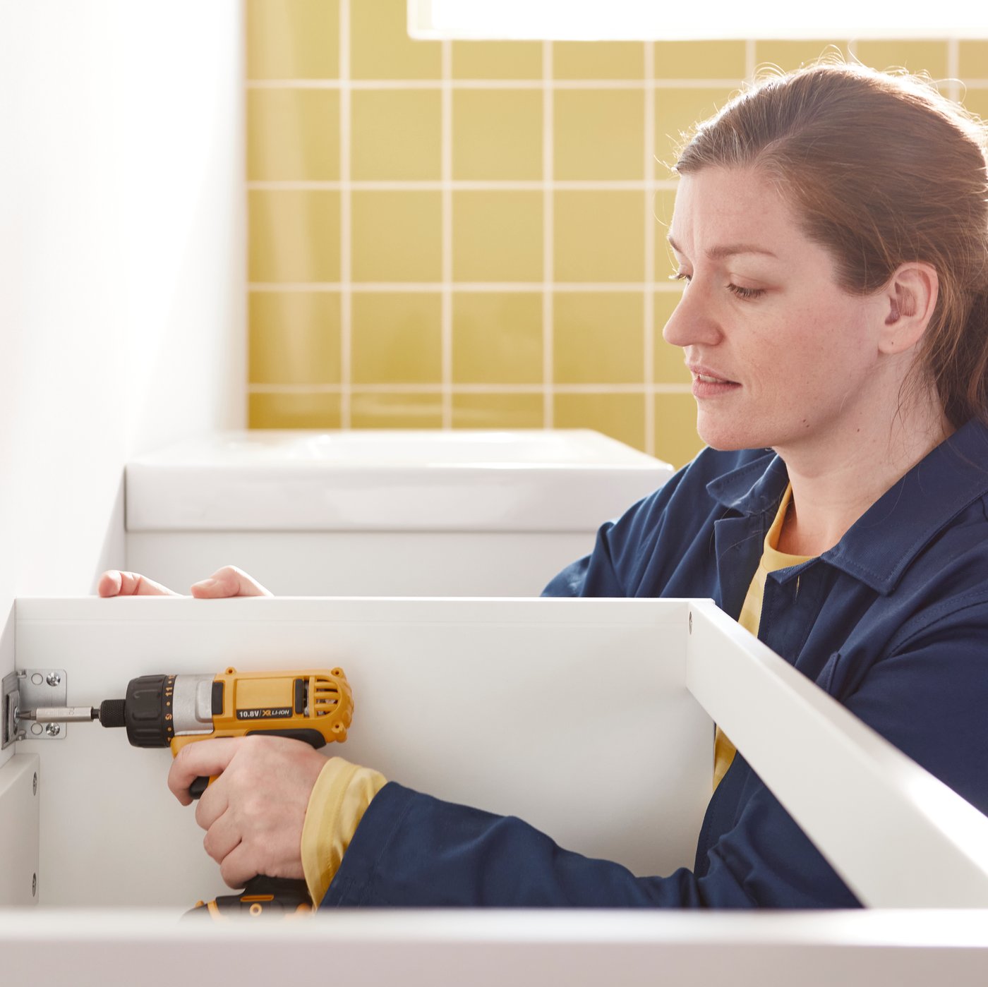 An IKEA co-worker is mounting a commode on to a bathroom wall with a screwdriver.