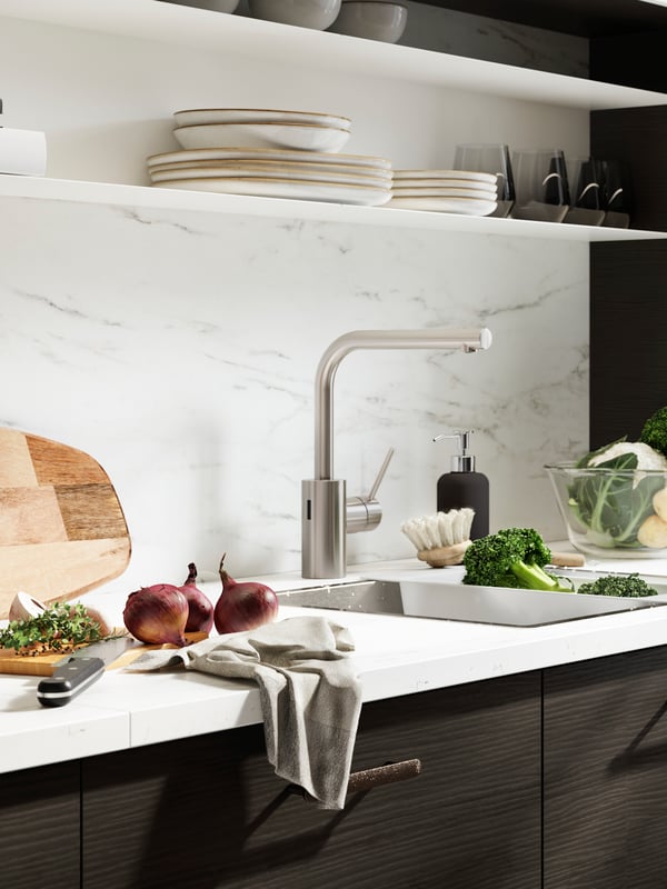 A kitchen with white worktop, brown fronts and white marbled wall panel. A cutting board and veggies beside the sink.