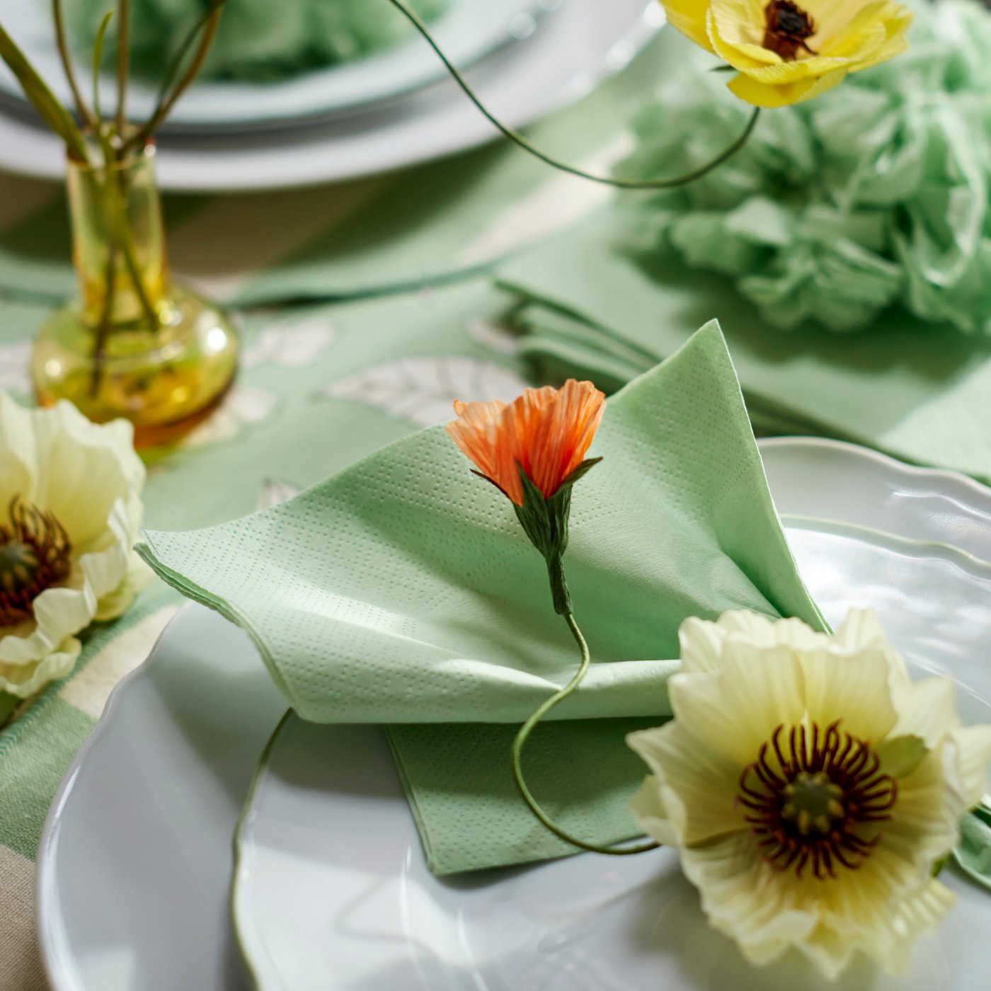 A green FANTASTISK paper napkin, on top of UPPLAGA side plate and plate, decorated with a SMYCKA artificial spray magnolia.