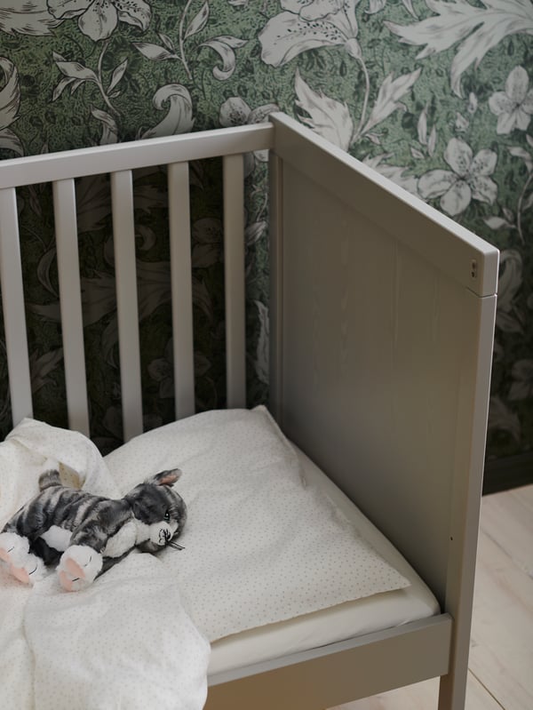 Kucing mainan lembut LILLEPLUTT terletak di atas katil bayi SUNDVIK berwarna kelabu dengan linen katil LENAST. Salah satu sisi katil bayi itu ditanggalkan.