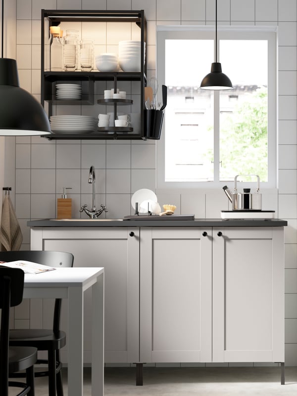 Vista frontal de un conjunto de muebles de cocina ENHET. El mueble superior de estructura negra es abierto y se ubica junto a una ventana, mientras que el mueble inferior, con lavaplatos incorporado, tiene una encimera gris oscura y puertas ENHET blancas con una cantería y perillas negras. 
