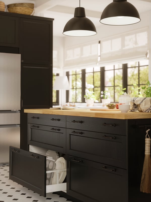 Un îlot de cuisine avec des façades LERHYTTAN teintées noires dont l’un des tiroirs est ouvert. Il y a des assiettes blanches à l’intérieur.