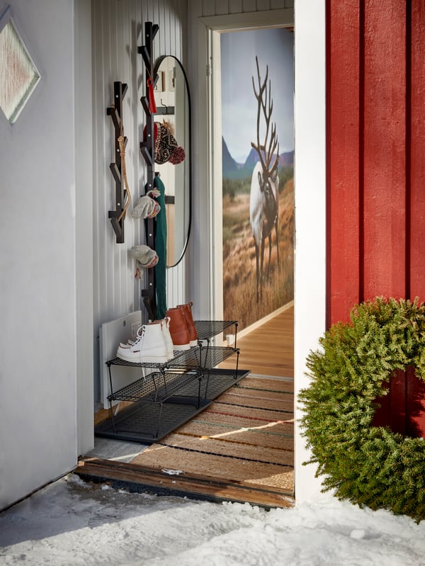 Öppen ytterdör på rött trähus där man ser ett trägolv, metallskoställ med vita skridskor, ytterkläder och hattar hängande på väggen samt en krans placerad på golvet vid dörren.