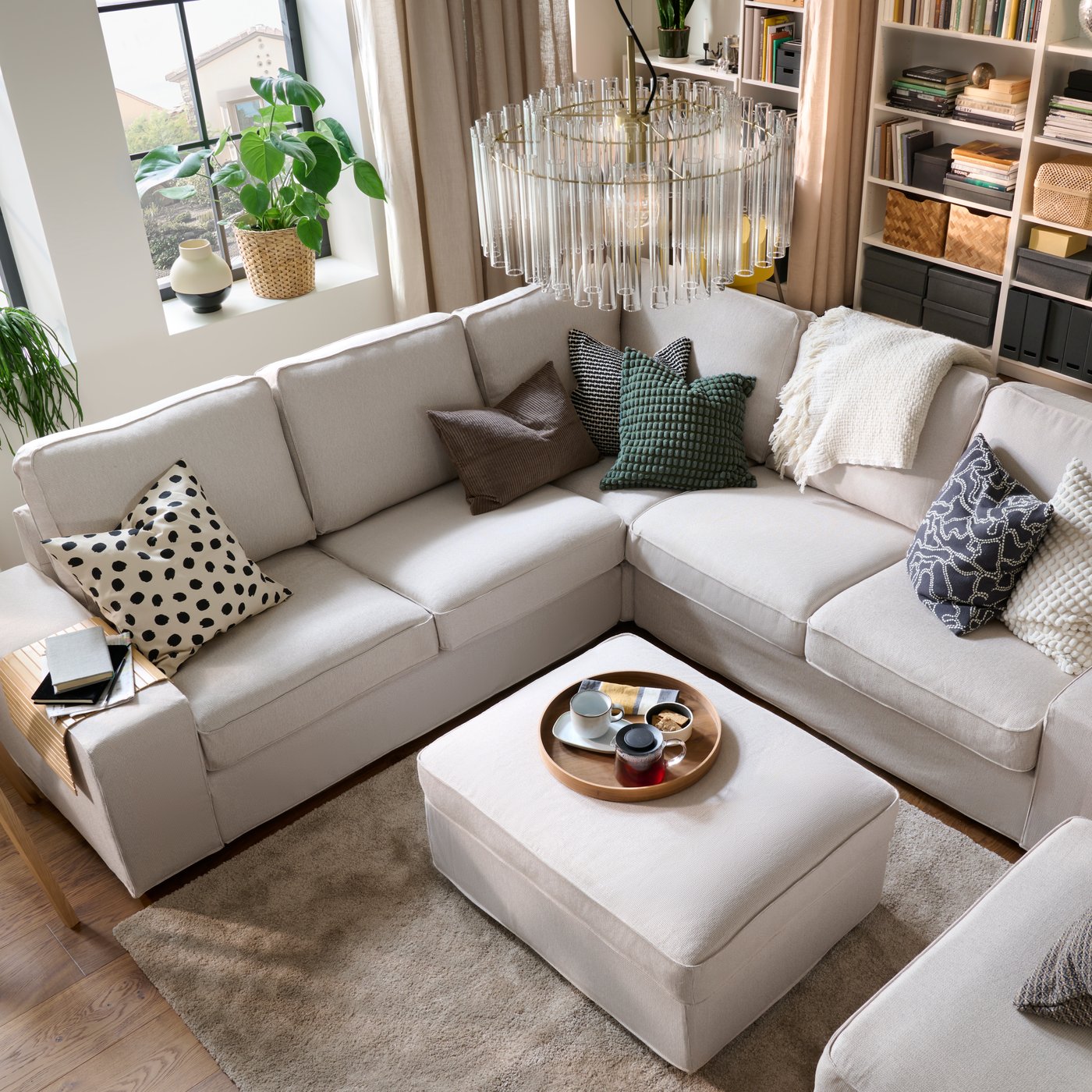 A light, bright living room with a KIVIK corner sofa and with colourful cushions, a throw and a centrally placed footstool.