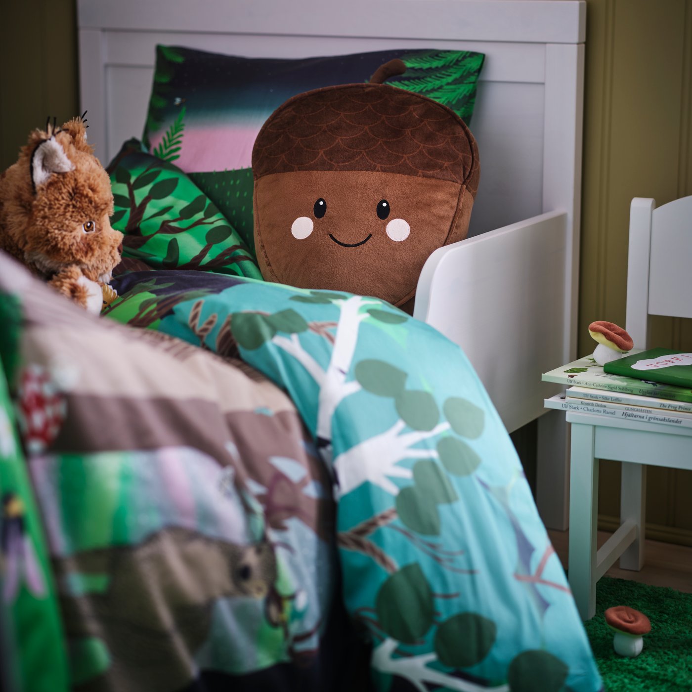 A SKOGSDUVA acorn-shaped cushion with pocket in a white bed with SKOGSDUVA duvet cover and pillowcase with a forest pattern.