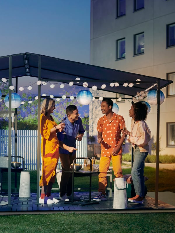 Two couples socialising outdoors at night in a GUNNÖN gazebo with RUNNEN outdoor floor decking and solar powered LED lamps.