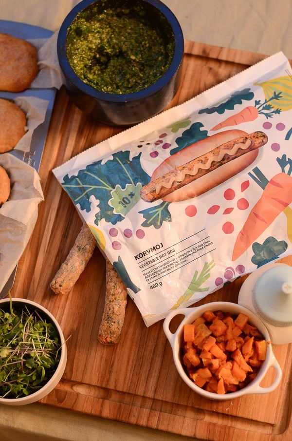 Ein Holzbrett mit Grissini, eine bunte Sandwichverpackung, Pesto, Microgreens, gewürfelte Süsskartoffeln und eine Flasche mit weisser Sauce, die an ein rustikales Picknick erinnern.
