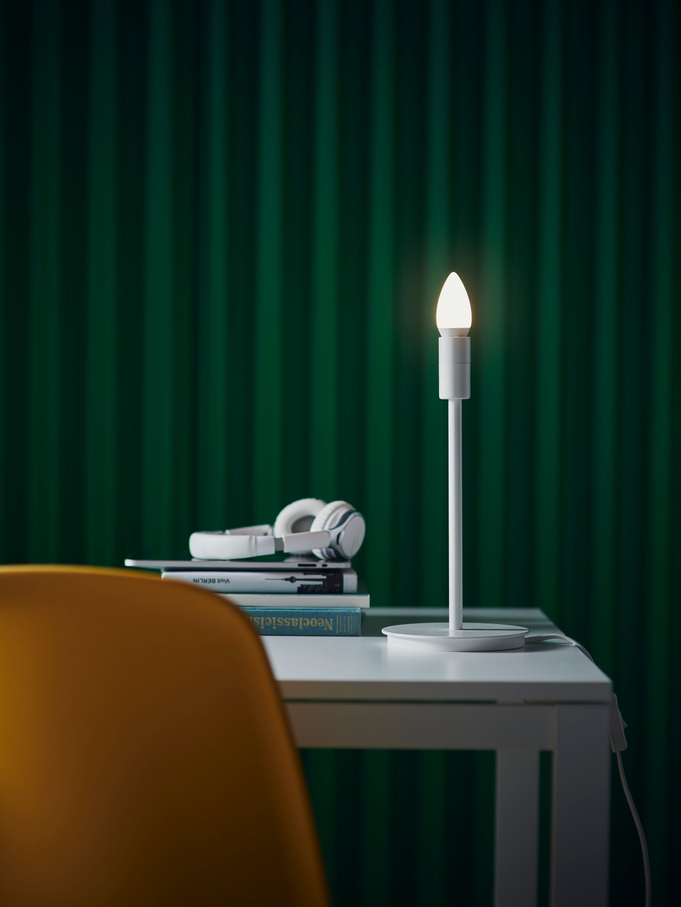 Une lampe de bureau blanche éclaire une table blanche sur laquelle sont posés une pile de livres et un casque audio blanc. Au premier plan se trouve une chaise orange, et le mur derrière elle est vert foncé.