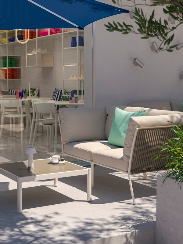 Et solrigt udendørs café-loungeområde med et hvidt/beige SEGERÖN udendørs samtalesæt med 2 siddepladser, en blå parasol og grønt.