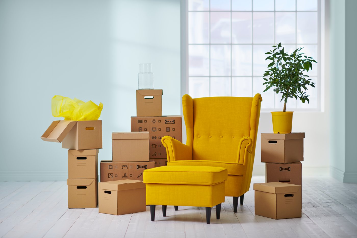 An empty house with a STRANDMON yellow armchair and a footstool, surrounded by DUNDERGUBBE cardboard moving boxes and PAPPIS cardboard boxes with lid and a plant.