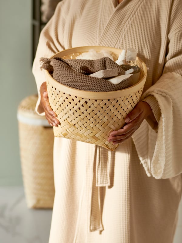 Une femme portant un peignoir beige tient un panier tressé SALUDING rempli de serviettes.
