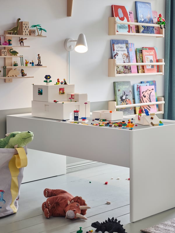 Na stole s úložným prostorem DUNDRA stojí stavebnice a krabice BYGGLEK LEGO®, na stěně za ním jsou knihy a další věci.