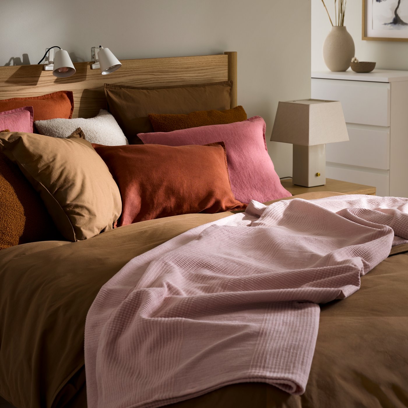 Ein Schlafzimmer mit einem Bett, das mit einer Kombination aus rosafarbenen und braunen Kissen dekoriert ist. Eine weiche rosafarbene Decke sorgt für eine gemütliche Atmosphäre.