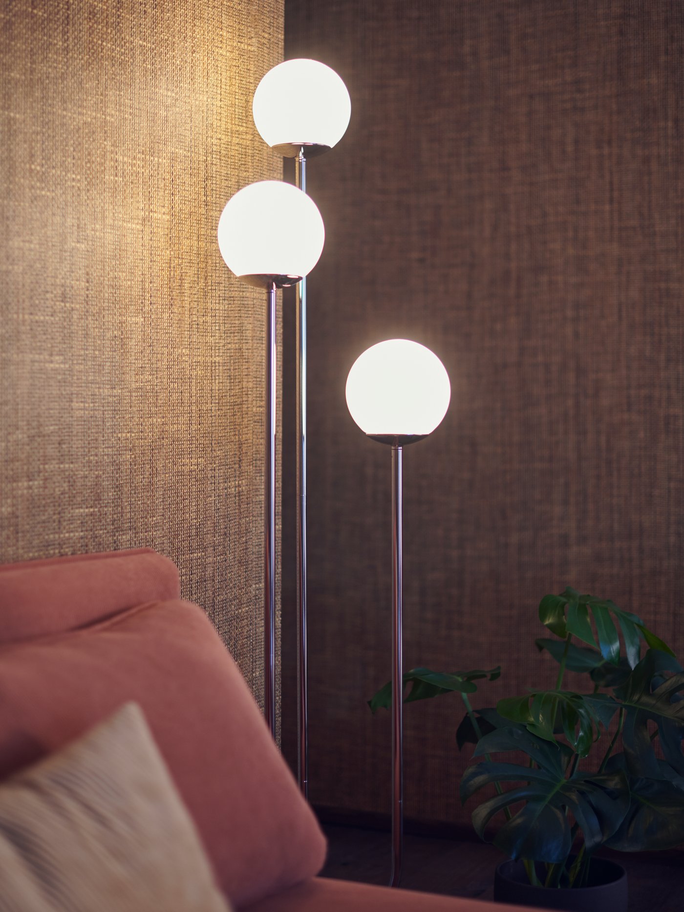 A SIMRISHAMN floor lamp with three bulbs giving off a warm glow against natural brown walls in the corner of a living room.
