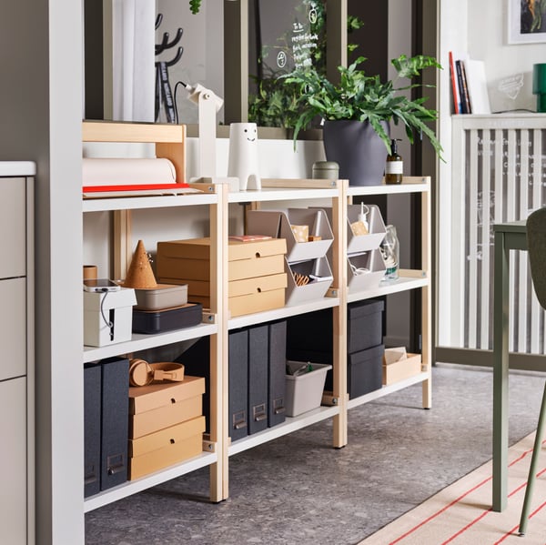 Open shelving units holding VATTENTRÅG boxes with lid, TJOG magazine files and other things stand under some windows.