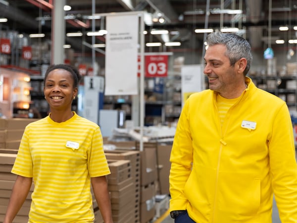 Twee IKEA medewerkers met lichtgele werkuniformen wandelen zijlings naast elkaar in een IKEA winkel.