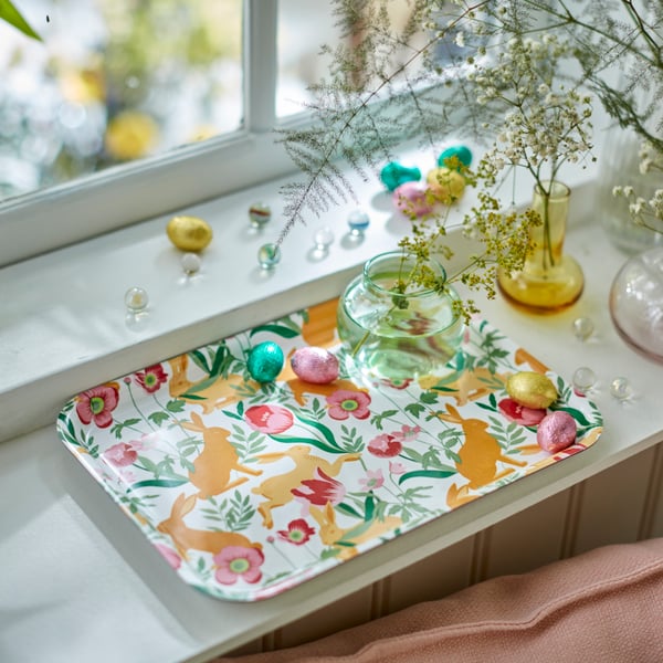A floral-patterned TJÄRLEK tray by a window, carrying VÅRKÄNSLA chocolate eggs and a vase with SMYCKA artificial sprays.