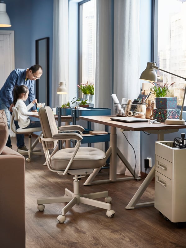 A man and a child in a room with IDÅSEN desks in ash veneer next to the windows, HEKTAR work lamps and beige office chairs.