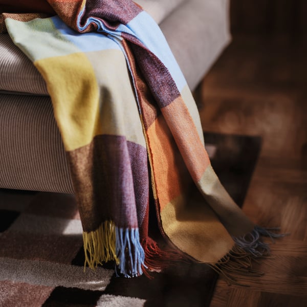 A close-up of a multicoloured FLIKHAGTORN throw with a large, checkered pattern softly draped over a beige sofa.