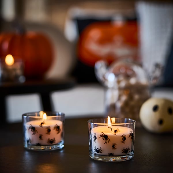 Two lit scented KUSTFYR candles with spider designs on glass placed side by side on a table.