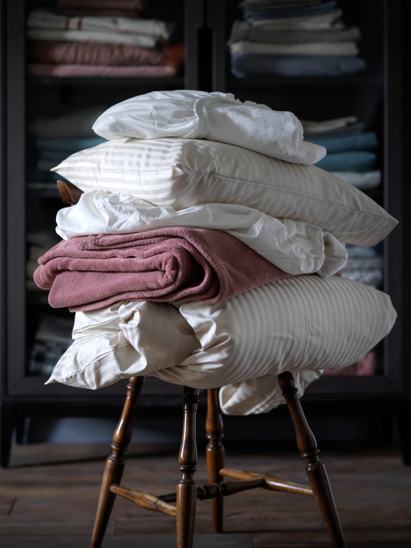 En hög med hopvikta sängkläder och kuddar i vitt och rosa, staplade på en trästol. I bakgrunden syns ett mörkt skåp med hyllor fyllda med textilier.