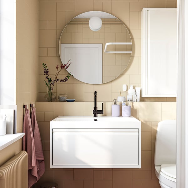 A white ÄNGSJÖN vanity with one drawer is mounted in a bathroom with light brown tiles below a round FAXÄLVEN mirror.