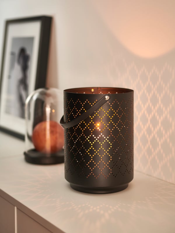 A black URSKILJA lantern for 1 block candle with a FENOMEN block candle lit inside, beside decorative items on a sideboard.