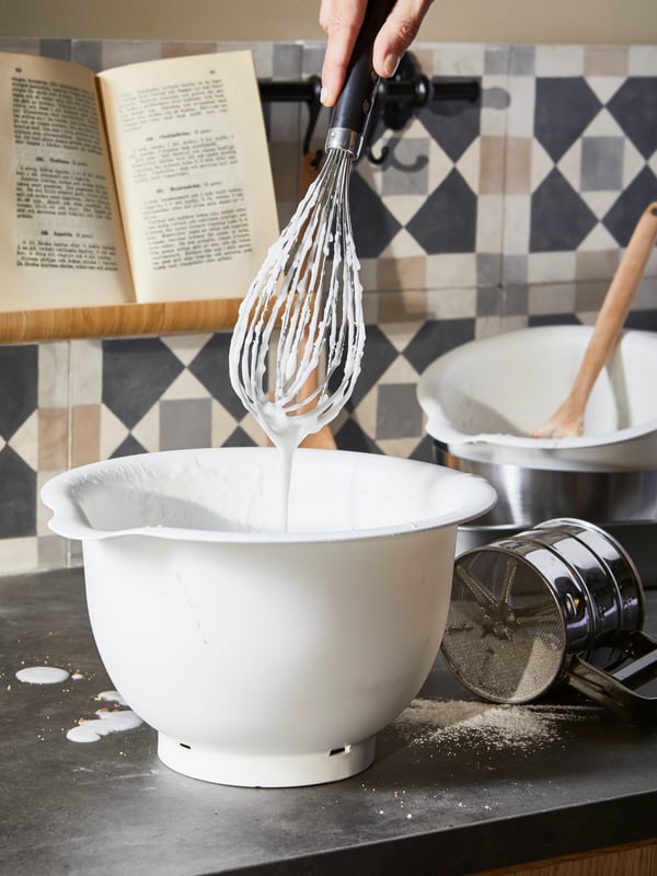 A hand is holding an IKEA 365+ balloon whisk over a white VISPAD bowling bowl standing on a kitchen counter.