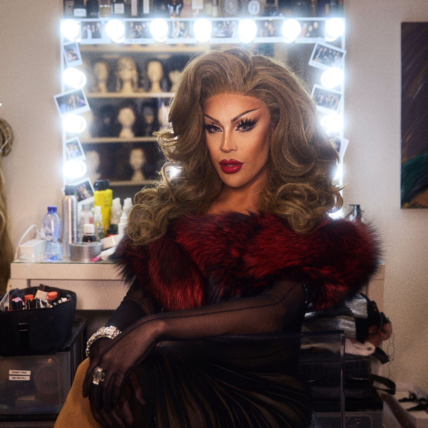 Envy Peru seduta nel suo camerino, con un trucco appariscente e travestita da donna. Lo specchio alle sue spalle proietta un bagliore glamour.