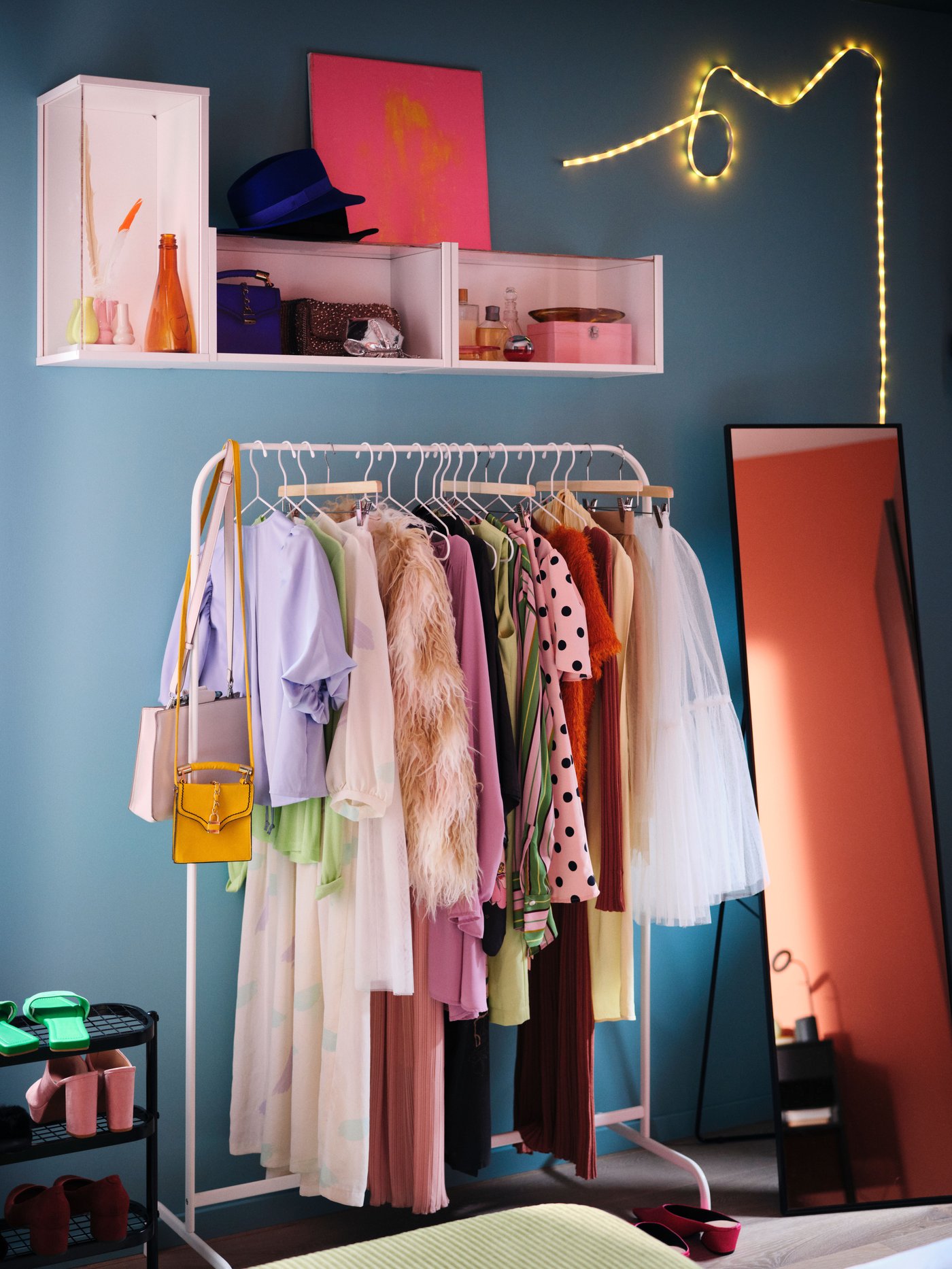 A blue dressing area with a white MULIG clothes rail holding colourful clothes beside a black KARMSUND standing mirror.