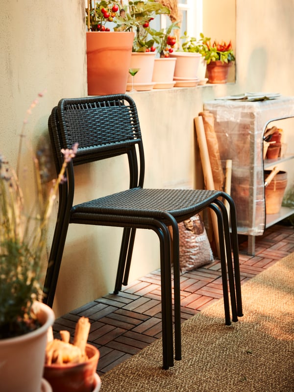 Tre sedie da esterno impilabili grigio scuro davanti a una parete gialla, un tappeto a tessitura piatta, uno scaffale e piante in vasi in terracotta - IKEA