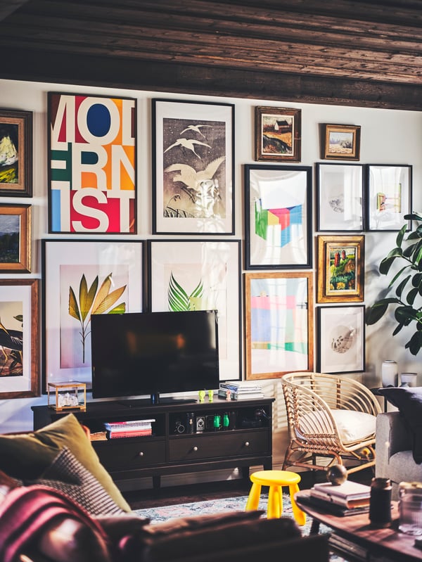 Una sala con un sillón de mimbre BUSKBO, una televisión, un mueble de televisión y una pared de galería con pósters BILD en marcos RIBBA y EDSBRUK. 