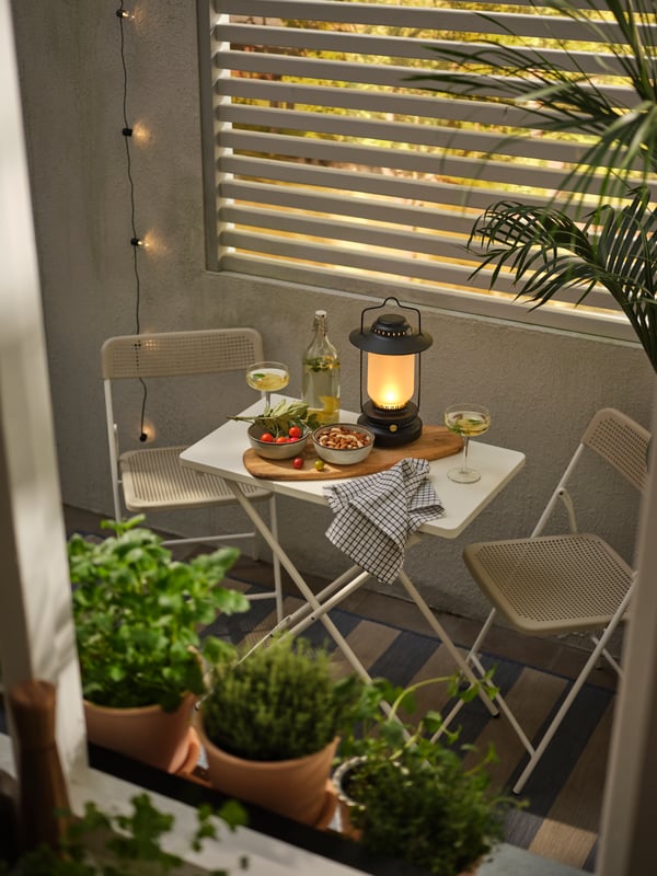 Un ensemble table et chaises TORPARÖ où se trouvent des boissons et en-cas, une planche à découper et une lampe de table STORHAGA, entourés de plantes.