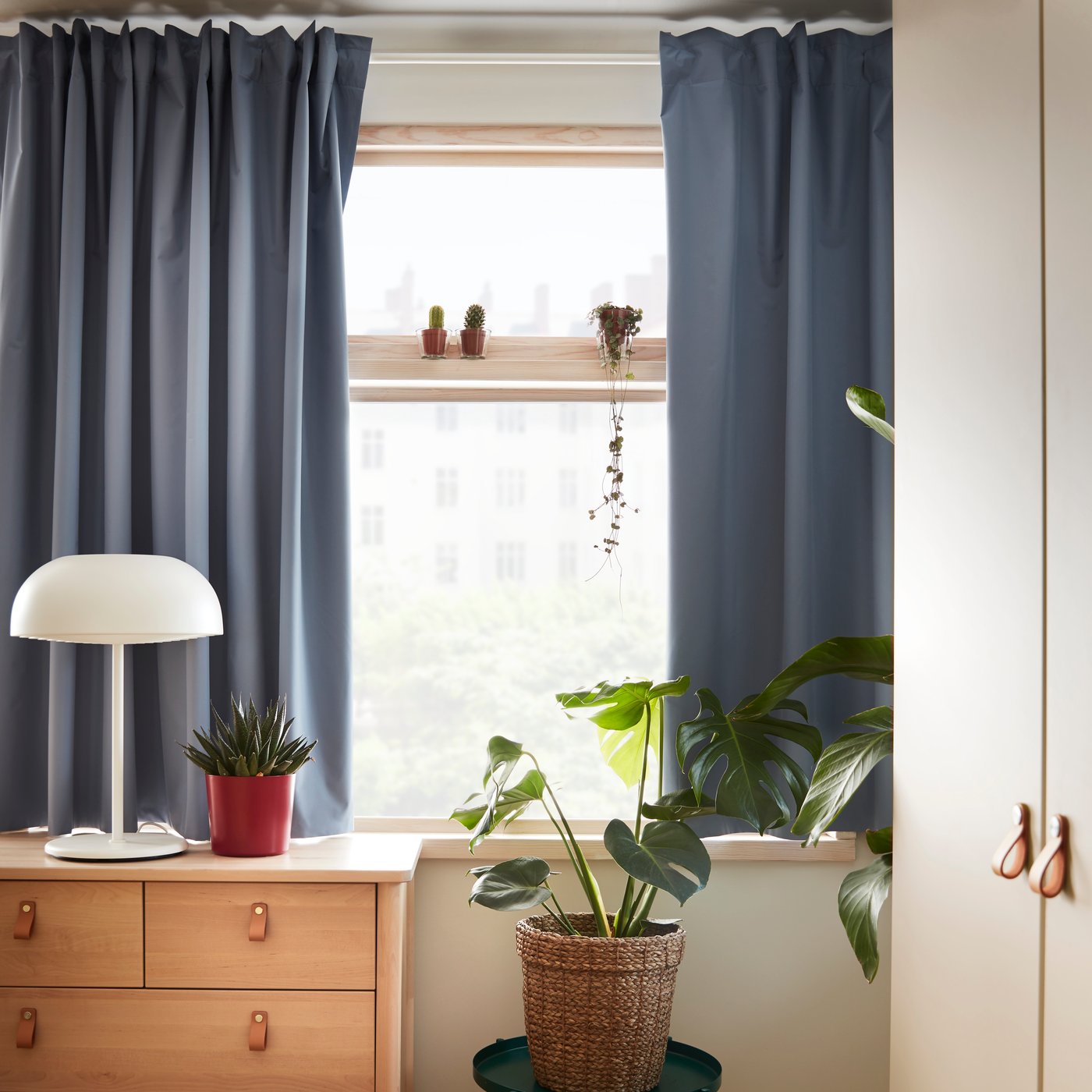 Vor einem Fenster steht eine braune Kommode auf der eine Tischlampe steht und eine Vase.
