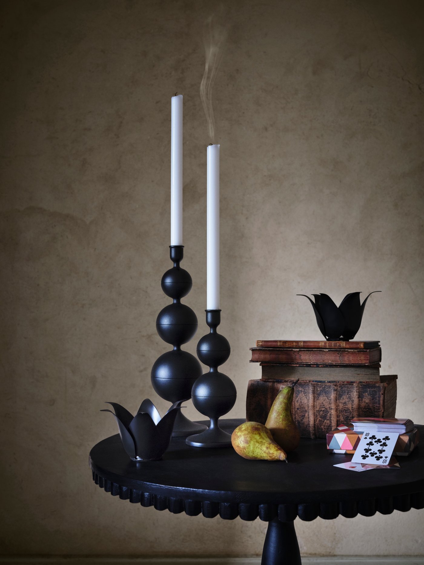 Two black DEKORERA candle sticks stand on a black table, with pears and books piled up next to it.